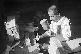 Historische schwarz-weiss Fotografie. Handwerker bearbeitet Holz mit Hammer und Meissel in Werkstatt, gestapelten Holzstücken auf der Werkbank.