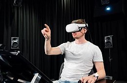 Mann mit weißem VR-Headset und Controller in einem dunklen Raum mit Lautsprechern, hebt eine Hand zum Interagieren.