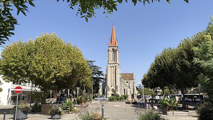 Platz im Genfer Vorort Lancy mit Bäumen, bepflanzten Behältern und einer Kirche mit rotem Turmspitzdach im Hintergrund