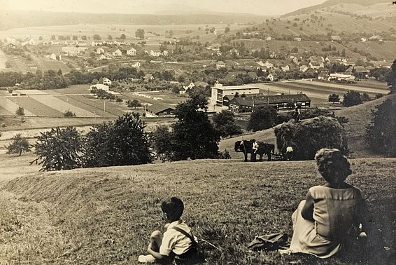 Blick auf Eglisau um 1930 mit Dorf, Feldern und Hügeln, zwei Personen sitzen im Vordergrund auf einer Wiese.