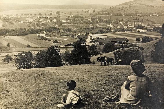 Blick auf Eglisau um 1930 mit Dorf, Feldern und Hügeln, zwei Personen sitzen im Vordergrund auf einer Wiese.