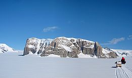 Forscher oder Forscherin auf Schlitten zieht ein Gerät über den schneebedeckten Starbuck-Gletscher vor einer felsigen, schneebedeckten Bergwand in der Antarktis.
