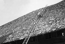 Historische schwarz-weiss Fotografie. Dachdecker klettert auf langer Leiter mit Holzschindeln gedecktes Dach eines Bauernhauses.