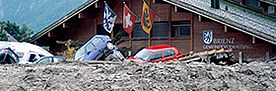 Vor der Gemeindeverwaltung in Brienz, einem grossen Holzhaus, türmen sich Schlamm- und Geröllmassen von einem Murgang. Verschiedene Autos liegen kreuz und quer davor. 