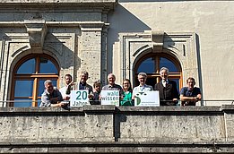 Gruppe von neun Personen auf einem Balkon vor einem historischen Gebäude mit großen Fenstern, einige halten Schilder mit Texten '20 Jahre' und 'wald wissen net'.