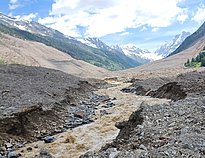 Schlammiger Gebirgsbach fließt durch ein von Geröll und Steinen bedecktes Tal mit schneebedeckten Bergen und bewaldeten Hängen im Hintergrund.