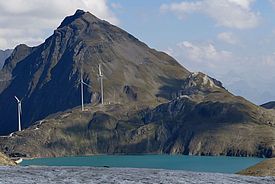 Windräder auf einem Bergkamm hinter einem schmelzenden Gletscher unter blauem Himmel mit Wolken