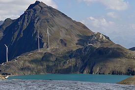 Windräder auf einem Bergkamm hinter einem schmelzenden Gletscher unter blauem Himmel mit Wolken