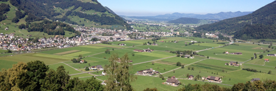 Blick auf ein Tal mit verstreuten Bauernhöfen, grünen Feldern und Bergen im Hintergrund bei klarem Himmel