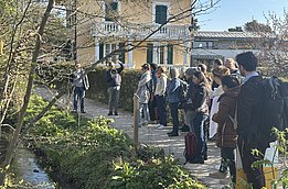 Gruppe von Menschen hört einer Person zu, die neben einem kleinen Bach vor einem gelben Haus spricht, Frühlingsbäume im Hintergrund.
