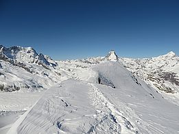 Schneebedeckter Grat mit einem Wander oder einer Wanderin, im Hintergrund das Matterhorn und weitere Alpenberge unter klarem Himmel
