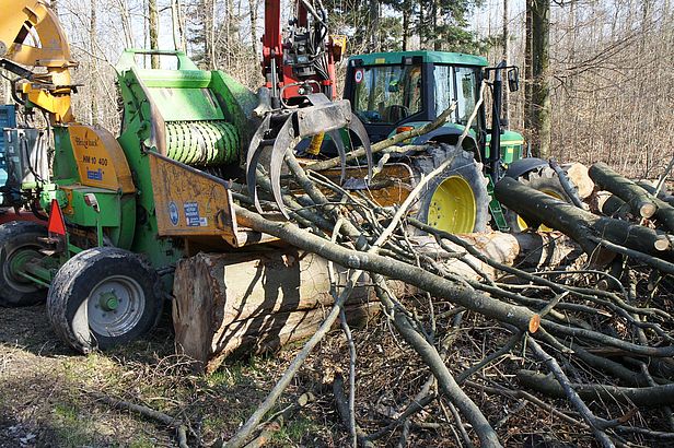 Holzernte: grüner Traktor mit Holzgreifer lädt Äste in eine gelbe Holzhäcksler-Maschine im Wald