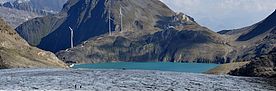 Griesgletscher mit Bergsee und Windrädern im Hintergrund unter blauem Himmel