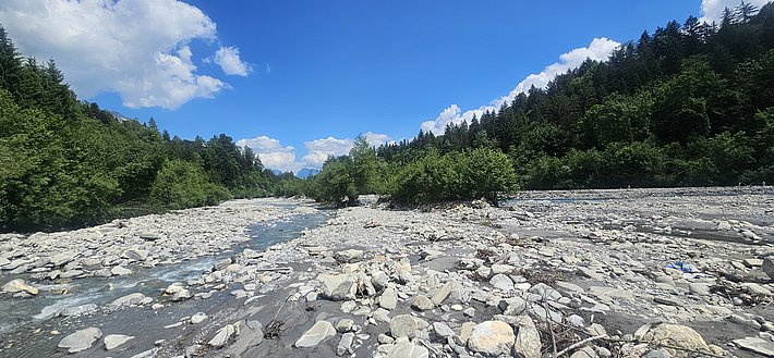 Fluss mit niedrigem Wasserstand, umgeben von Kieselsteinen und bewaldeten Hügeln unter blauem Himmel mit Wolken