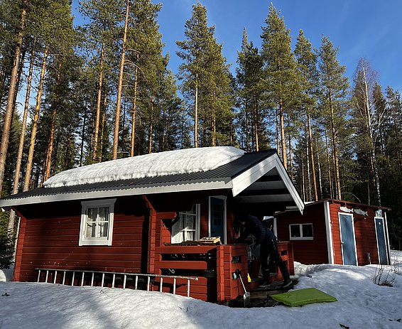 Rotes Holzhaus mit schneebedecktem Dach in einem verschneiten Wald mit hohen Kiefern, Person auf der Veranda