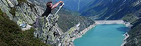 Person arbeitet mit Gerät an felsigem Berghang mit Blick auf türkisfarbenen Stausee in Tal