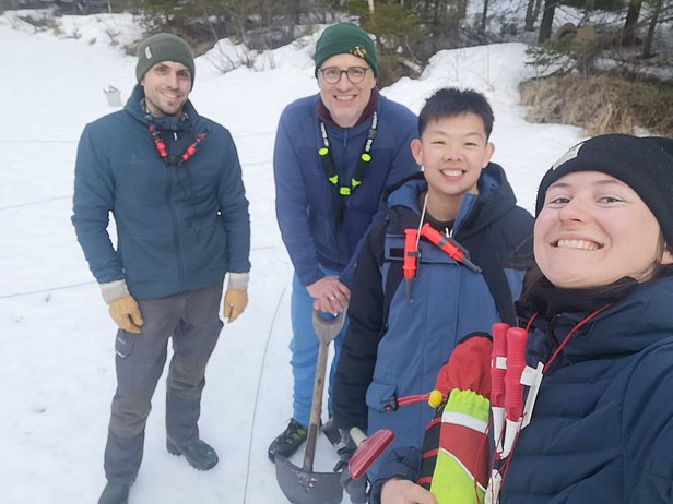 Vier Personen in Winterkleidung stehen lachend im Schnee, eine hält eine Schaufel, Wald im Hintergrund