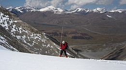 Person in roter Jacke und Helm misst Schneetiefe in verschneitem Gebirgstal mit schneebedeckten Gipfeln im Hintergrund